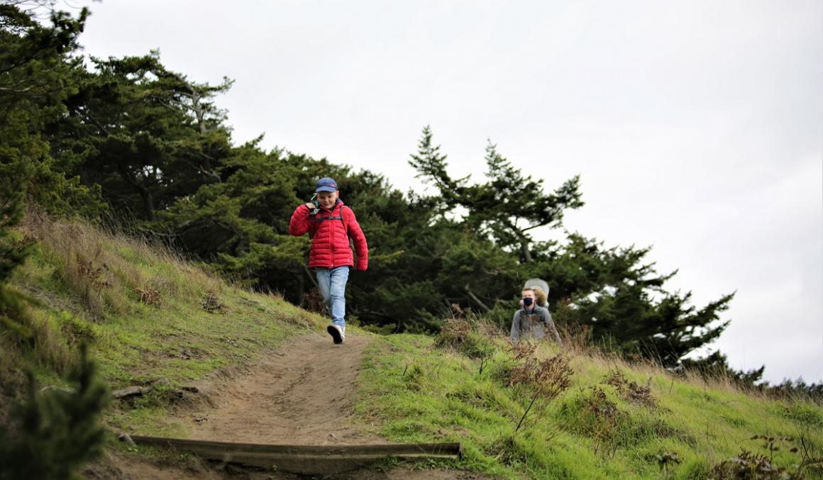 Boy in red jacket followed by father with baby in carrier on hiking trail in Washington mountains