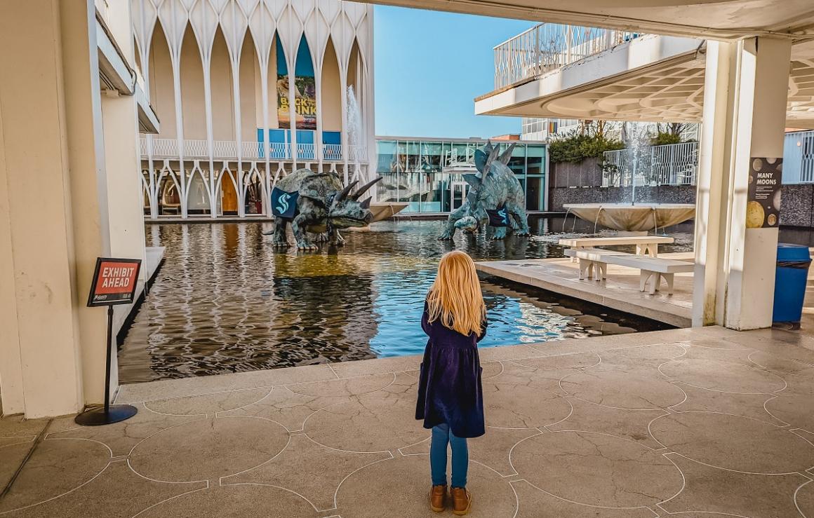 Dinosaur statues in front of Pacific Science Center wear Seattle Kraken hockey team gear as a girl looks on