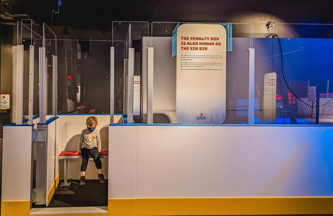 A young boy of about 2 wears a mask and sits in model penalty box or "sin bin" at new exhibit Hockey: Faster Than Ever at Seattle's Pacific Science Center