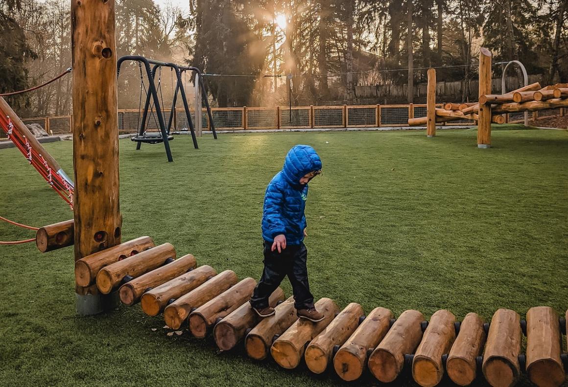 Westside Park in Redmond young boy in blue jacket walking on log bridge among natural wood play structures new playground