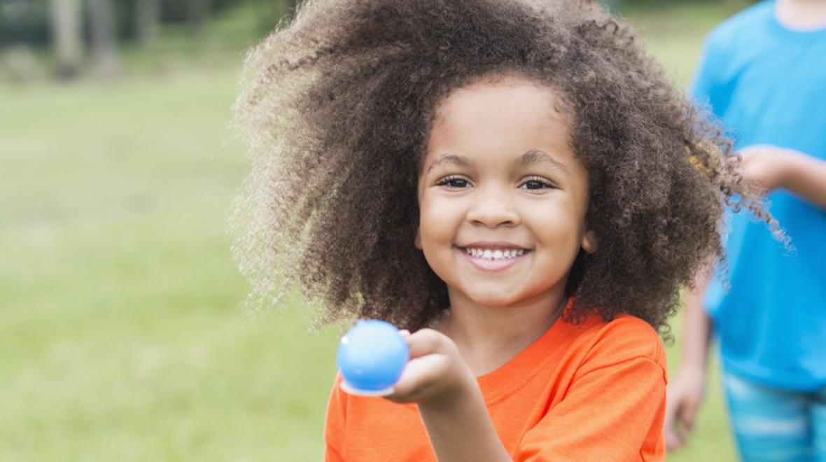 little kid holding a blue egg on a spoon outdoors