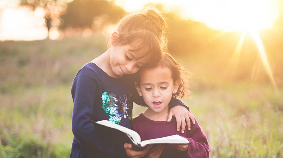 two sisters standing in a field holding a book with the sun behind them