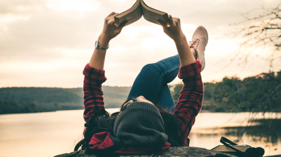 Teenager lying on back holding up  book, reading. Water in the background